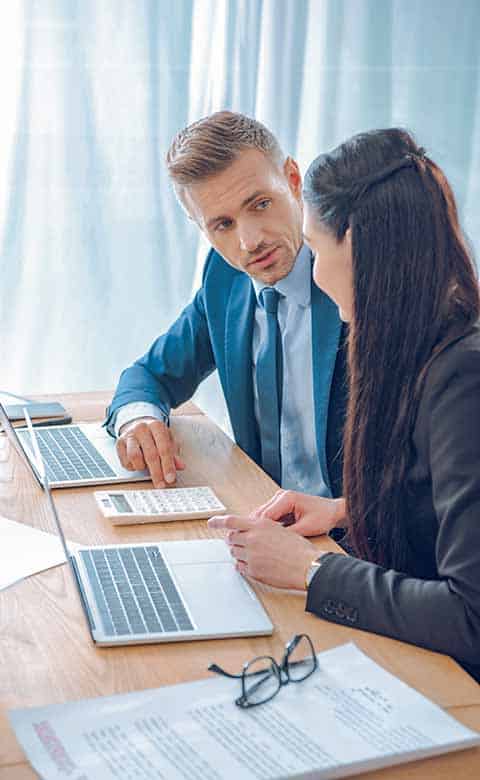 man & woman sitting at desk, in front of laptops using a calculator